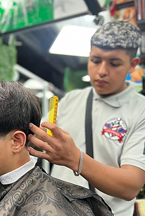 Barbero con playera del logo de Los Panas cortando con peine amarillo a un cliente con capa estampada