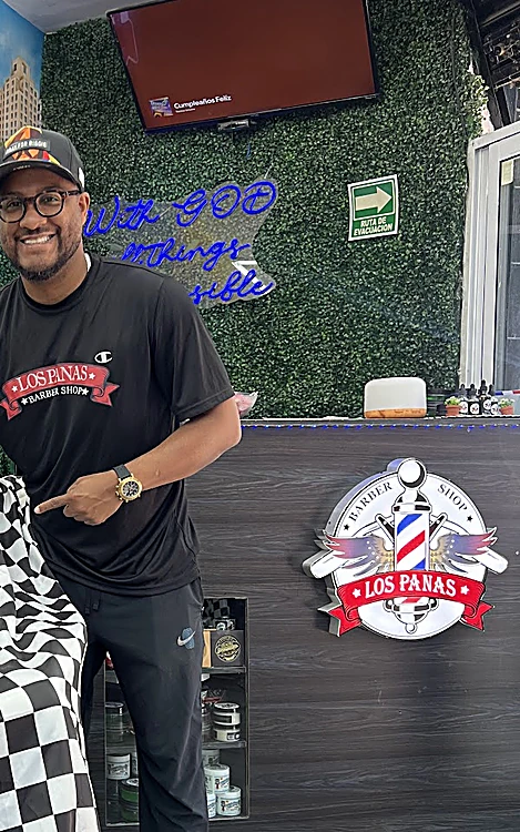 Barbero de Los Panas sonriendo junto al mostrador de madera negra con el logo de la barbería y un neón azul en el muro de pasto