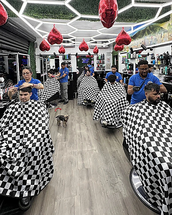 El salón de Los Panas a lo largo: fila de sillones con capas a cuadros blanco y negro, barberos con playera azul trabajando y piso de madera